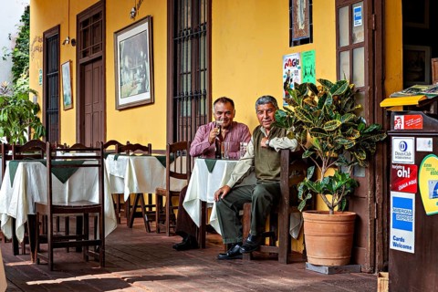 Two men sit comfortably outdoors at a table in front of a yellow building and enjoy the atmosphere,