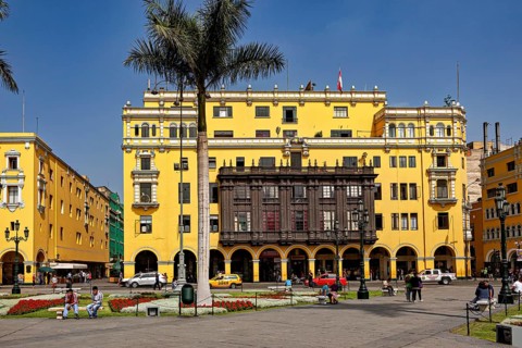 Wide square with yellow colonial buildings and palm trees surrounded by people and cars, The