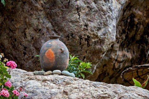 An ancient clay vase on stones in front of a rocky landscape with flowers in the foreground, a clay