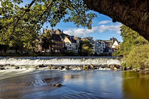 City scene with river and waterfall, surrounded by old buildings and trees under a blue sky, Das
