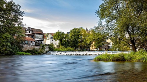 River landscape with historic buildings and trees, quiet atmosphere under blue skies, Das Wehr der