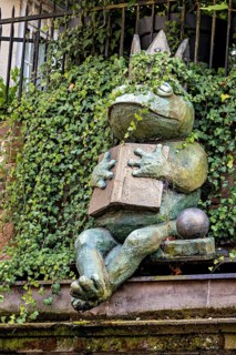 A frog sculpture with crown and book, surrounded by ivy, creates an enchanted atmosphere, The Frog