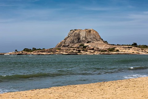 Tranquil coastal landscape with a large rock in the sea, surrounded by sandy beach and blue sky,