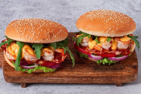 Two shrimp burgers and buns with sesame seeds, bacon, lettuce, tomatoes and onions, On a wooden