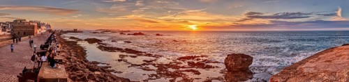 Panorama of a coastal promenade at sunset with rocks in the sea and a bright orange sky, Morocco,