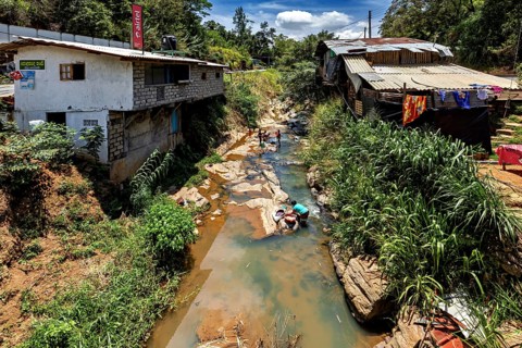 A river flows through a village, people wash clothes surrounded by nature and simple buildings,
