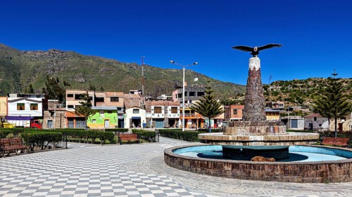 A public square with fountain and bird statue, surrounded by colorful houses and mountains, the