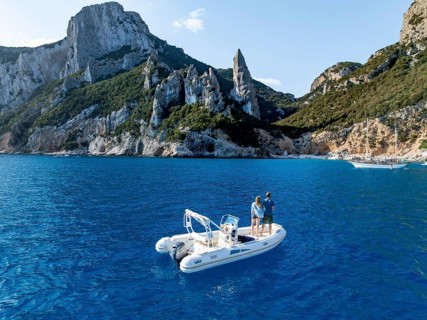 Couple on motorboat off picturesque rocky coast, cliffs with L'Aguglia pinnacle, blue sea and Cala