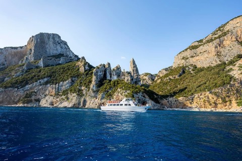 Excursion boat off picturesque rocky coast, cliffs with caves in morning light, L'Aguglia rock pin,