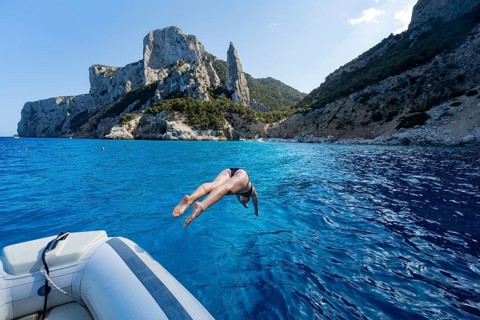 Young woman jumping from a boat into the water, picturesque rocky coast, cliffs with L'Aguglia rock