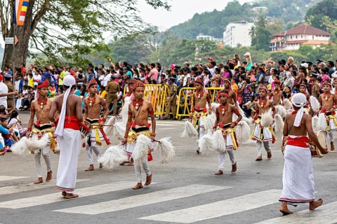 Children move across a street in traditional costumes, spectators watch, The Perahera Parade in