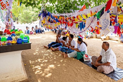 People sitting praying under colorful flags in the shade of a tree on sandy ground, pilgrims to the