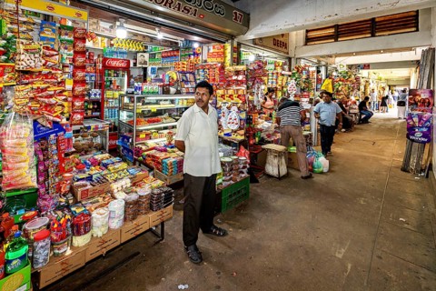 A man stands in a well-stocked shop with numerous colorful foods and drinks, dealers in the market