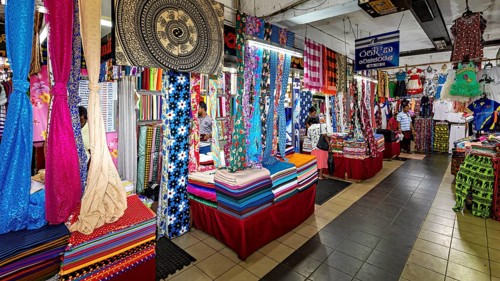 Various colorful fabrics and textiles are on display in a busy market hall, dealers in the market