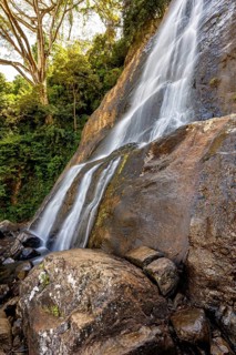 The clear water of a waterfall flows over large rocks, surrounded by green vegetation and tall