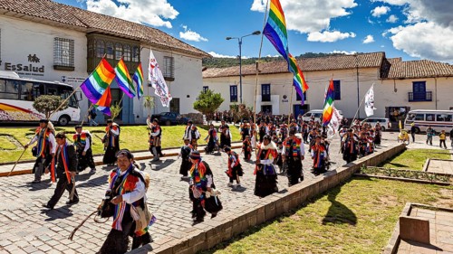 People in a colorful parade with rainbow flags walk along under a sunny sky, traditional parade of