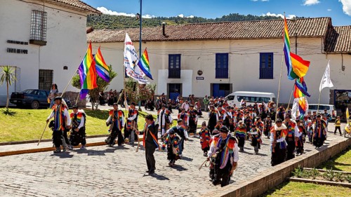 People with colorful flags march through a street with historic buildings under blue skies,
