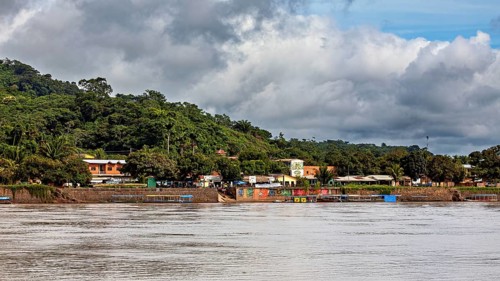 A quiet riverside village with houses in front of a wooded hill under cloudy skies, the city of