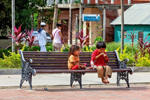 Two children sit on an outdoor bench while they eat. The surrounding area is green and peaceful,