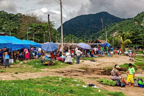 Outdoor market in front of a mountain landscape, with stalls and people under blue tarpaulins, The