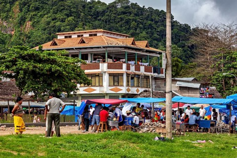 Large house in the background of a busy market square with people and mountains in the distance,