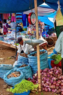 Market scene with vegetable stands, bags full of potatoes and onions. A scale hangs over the stand,