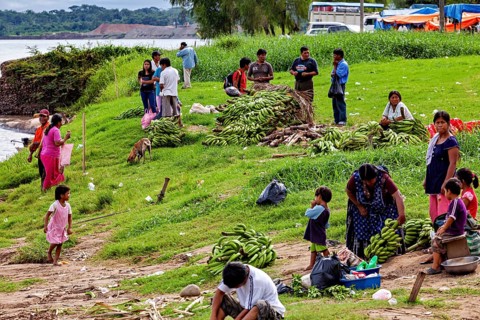 People sell bananas on a riverbank in a rural area. The scene depicts a communal and bustling