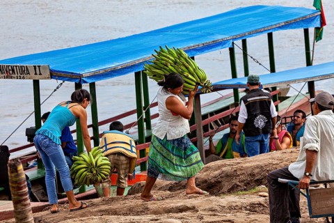 Two woman carry bundles of bananas to a riverbank boat as people watch, woman unload bananas from a