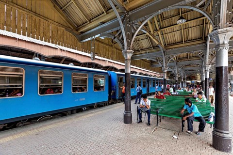 People sitting on green benches in the train station, blue train on the platform, covered