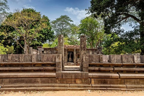 Stone gate marking the entrance to an ancient structure under a clear sky, surrounded by trees, The