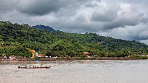 A boat moves on a river in front of a wooded hill and under a cloudy sky, people on boats near