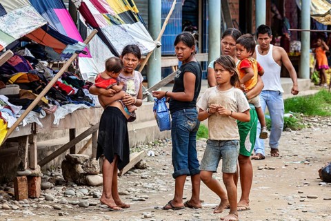 Group of people, including woman and children, stand at a market with colorful stalls, poverty in