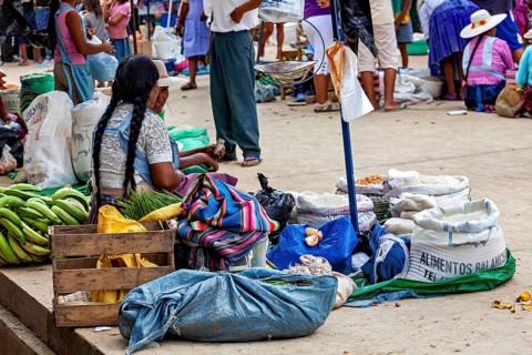 A woman sits in a market surrounded by sacks and bananas and observes the surrounding area, The