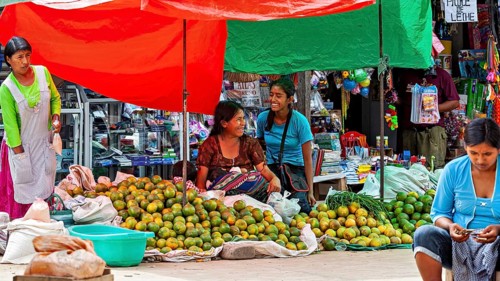 Two laughing woman sit in front of market stalls full of mangoes in a lively atmosphere, The