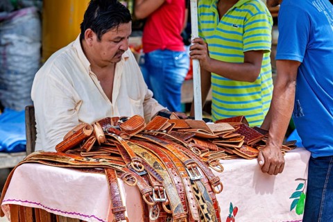 A man sells leather belts at a market stall surrounded by customers, The Rurrenabaque market in