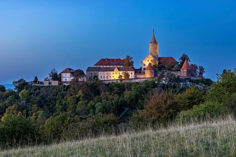 An illuminated castle on a hill at dusk, surrounded by shady trees, The Leuchtenburg near Kahla im
