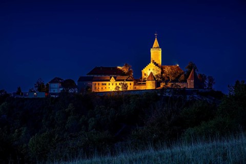 An illuminated castle is perched on a hill, surrounded by dark countryside and a deep blue night