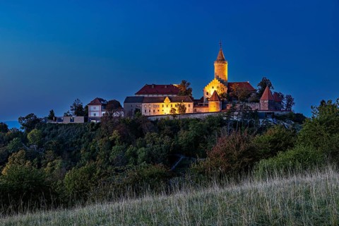 A castle on a wooded hill, illuminated at dusk with dark skies, The Leuchtenburg near Kahla im