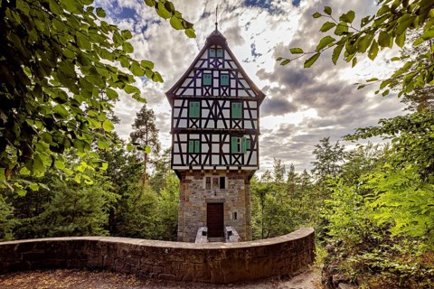 Half-timbered house in the forest surrounded by leaves, under a cloudy sky, with a curved stone