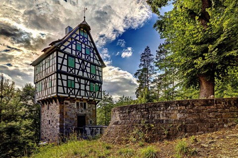 Half-timbered house on a stone base in the forest, surrounded by trees and a stone wall, under a