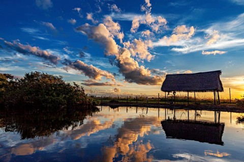 A thatched hut reflected in the water at sunset with dramatic cloud formations, The pampas swamps