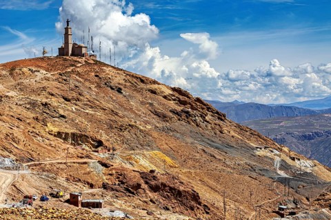 Rough mountain terrain with sparse vegetation and a transmission mast on the summit under a cloudy