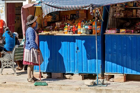 Woman in traditional dress sweeps in front of a blue market stall full of colourful bottles and