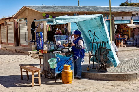 Street snack bar under a blue sunshade, woman serving customers next to a table and gas canister in