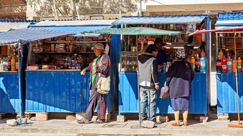 Street market with a blue stall, people buy colourful bottles and products under a blue sky, small
