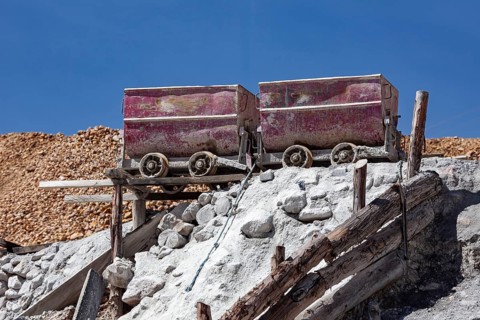 Two rusty mine cars on a wooden scaffold, surrounded by piles of rocks in a sunny mining