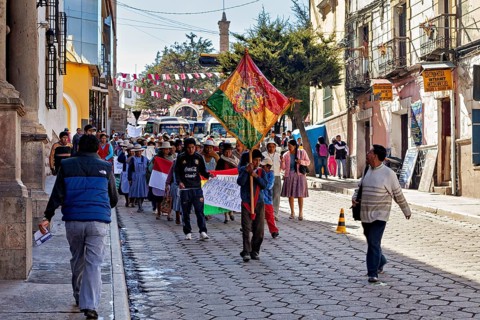 People demonstrate with flags and banners on a cobbled street between historic buildings,