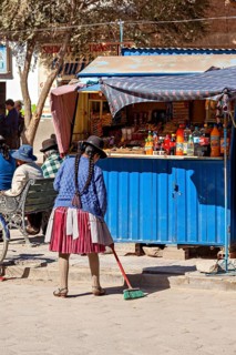 Woman in traditional dress sweeping in front of a blue stall on a busy street in the sunshine,