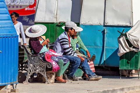 People with hats sitting on a bench in the sun at a market stall, people of the city of Uyuni in