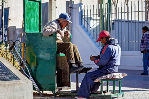 Shoe shiner concentrates on a man sitting on a green bench outdoors, show shiner in the city of